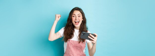 Excited beautful girl winning on mobile phone holding smartphone and shouting yes with joyful face a