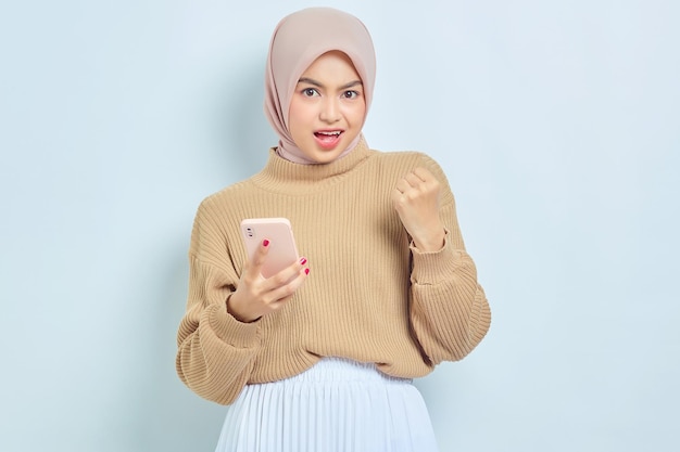 Excited beautiful asian muslim woman in brown sweater and hijab\
using a mobile phone celebrating success getting good news isolated\
over white background people religious lifestyle concept