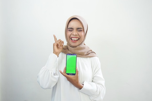 Excited beautiful Asian muslim woman showing green screen mobile phone isolated on white