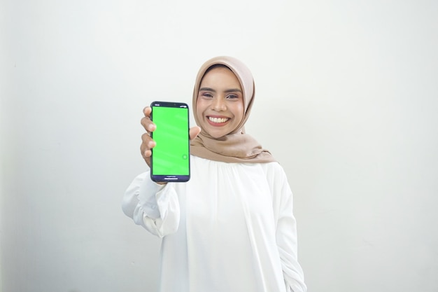 Возбужденная красивая азиатская мусульманка показывает мобильный телефон с зеленым экраном, изолированный на белом