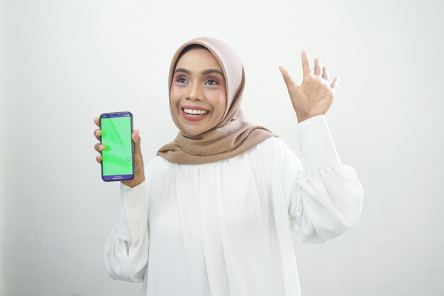 Excited beautiful Asian muslim woman showing green screen mobile phone isolated on white