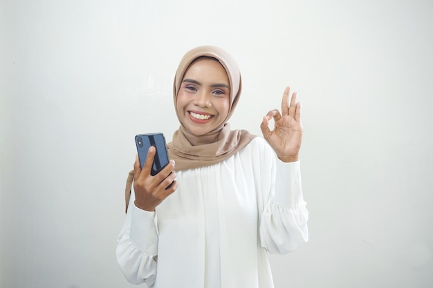 Возбужденная красивая азиатская мусульманка показывает мобильный телефон на белом фоне