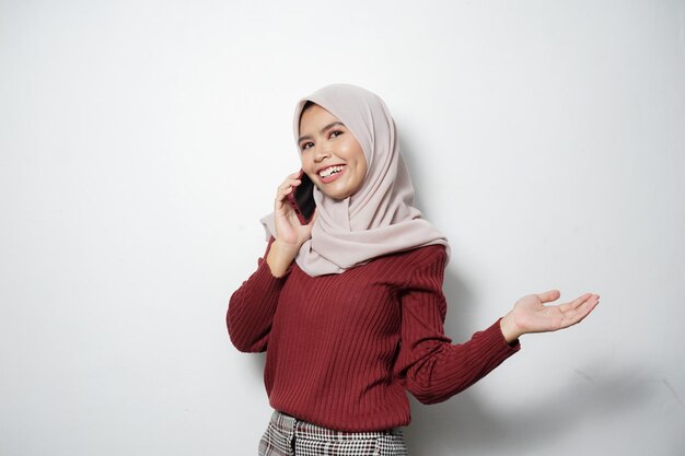 Excited beautiful Asian woman in maroon sweater and hijab using a mobile phone celebrating success getting good news isolated over white background