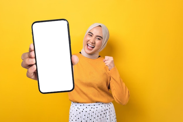 Excited beautiful Asian woman wearing hijab holding smartphone with blank white screen and clenching fist celebrating victory isolated on yellow background mockup