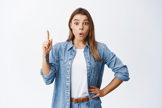 Excited beautiful girl making good point recommending deal pointing finger up or pitching idea making eureka gesture standing against white background