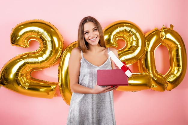 Excited beautiful woman opening gift box in front of new year 2020 balloons isolated over pink