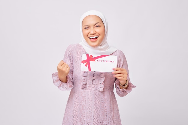 Excited beautiful young Asian Muslim woman wearing hijab and purple dress holding gift voucher certificate and celebrating winner isolated on white studio background