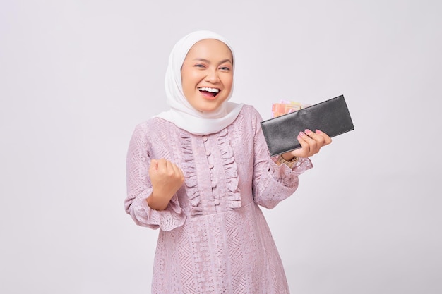 Excited beautiful young Asian Muslim woman wearing hijab and purple dress holding wallet of full cash money and celebrating succes isolated on white studio background