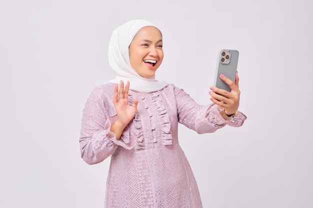 Excited beautiful young Asian Muslim woman wearing hijab and purple dress using smartphone making video call or taking selfie isolated on white studio background