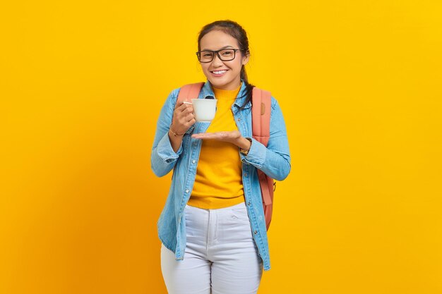 Excited beautiful young asian woman student in denim clothes with backpack while showing cup of coffee with palms isolated on yellow background