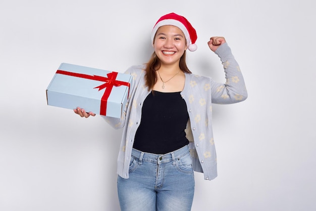 Взволнованная красивая молодая азиатка в шляпе Санта-Клауса, показывающая подарочную коробку и празднующая удачу на белом фоне студии Merry Christmas Concept