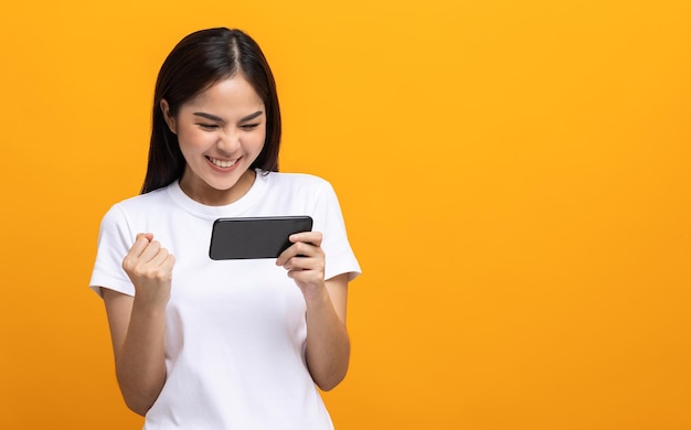 Excited Beautiful young asian women play mobile game and standing on isolated yellow background. Playing game on smartphone winning victory moment. Very enjoy and fun relax time