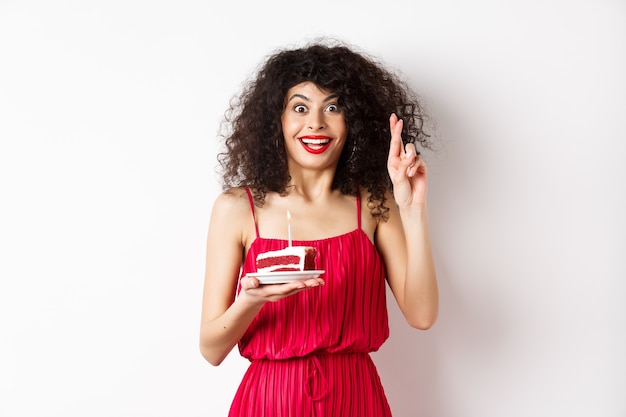 Ragazza di compleanno eccitata in vestito rosso, dita incrociate mentre esprime il desiderio e soffiando la candela sulla torta di compleanno, sorridendo felice, sfondo bianco.