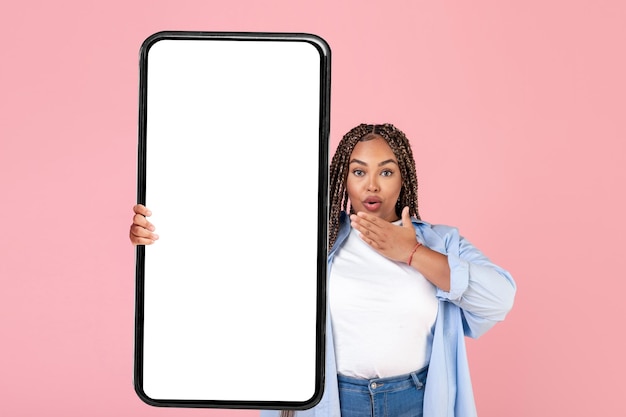 ピンクの背景に大きなスマートフォンの空の画面を保持している興奮した黒人女性