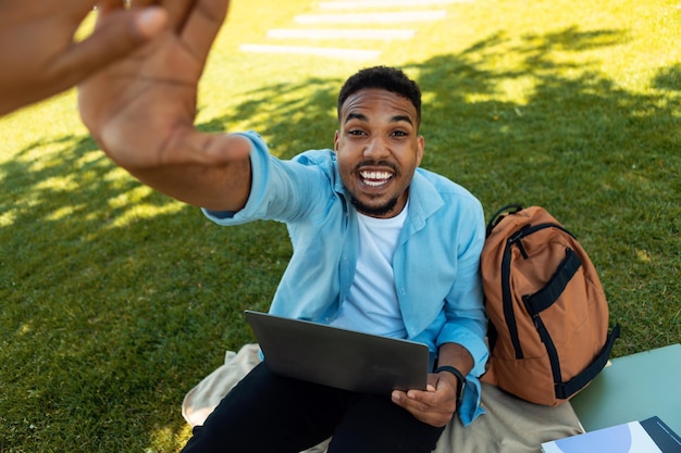 Excited black male student giving high five to friend while using laptop computer sitting on grass