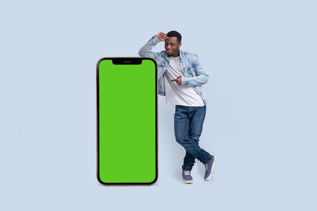 Excited black man standing and pointing Big smartphone with blank green screen demonstrating copy space for app or website design standing over blue studio background mockup image