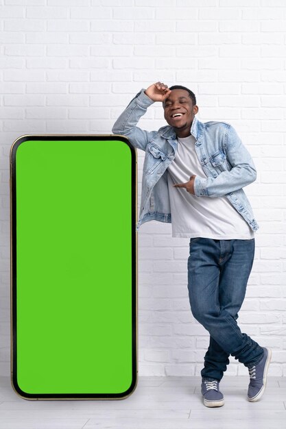 Excited black man standing and pointing Big smartphone with blank green screen demonstrating copy space for app or website design standing over white brick background mockup image