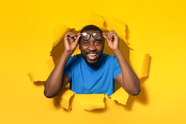 Excited black man taking off eyeglasses and smiling at camera\
standing in torn yellow paper hole