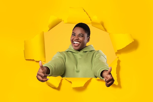 Excited black plus size woman gesturing thumbs up stretching arms to camera through hole in torn yellow paper