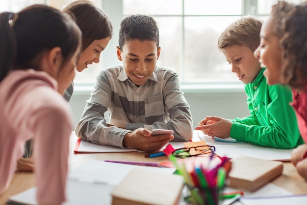 Photo excited black schoolboy using phone with classmates in modern classroom