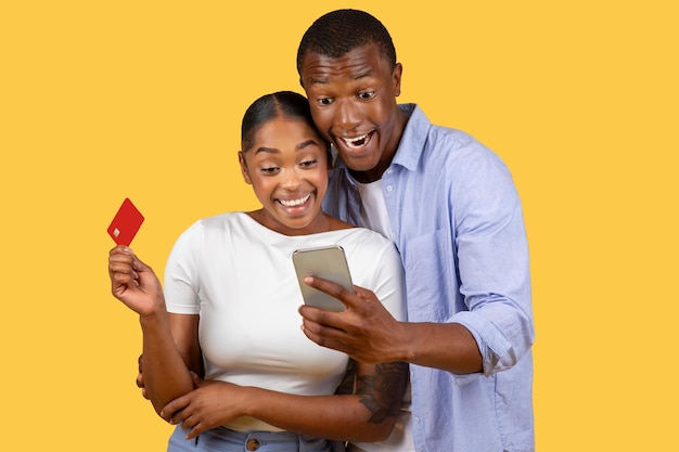 Excited black spouses shop online with phone and card discovering great deals on yellow background