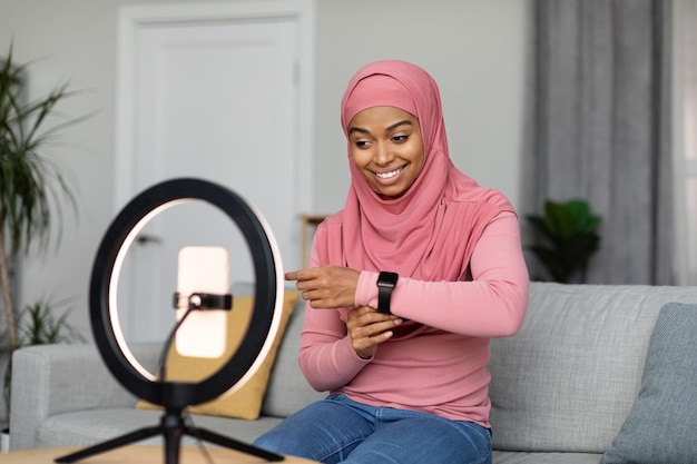 Foto eccitata donna di colore in hijab che fa una panoramica dei dispositivi e mostra video di registrazione di orologi intelligenti sul cellulare a casa