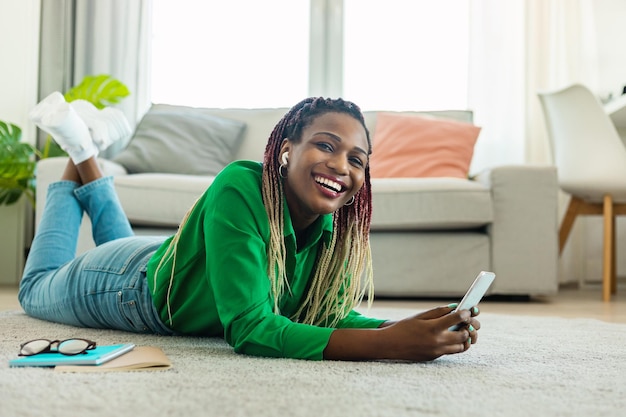 Возбужденная чернокожая женщина слушает музыку в наушниках с помощью смартфона, лежа дома на полу и улыбаясь в камеру
