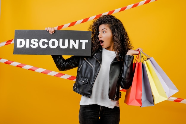 Excited black woman with discount sign and colorful shopping bags isolated over yellow with signal tape