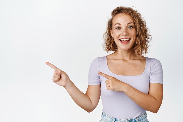 Excited blond caucasian girl pointing fingers left, smiling and looking happy, showing announcement, standing over white background