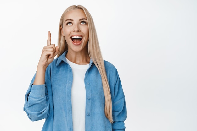 Ragazza bionda eccitata che sembra affascinata che punta il dito verso l'alto controllando l'annuncio di vendita sopra in piedi su sfondo bianco