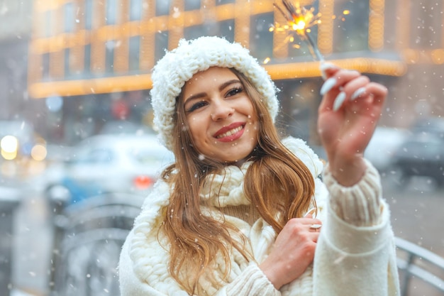 Eccitata donna bionda in abbigliamento invernale che si diverte con le stelle filanti durante la nevicata