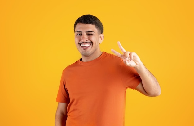 Excited brazilian man making peace sign with hand and winking at camera isolated on orange