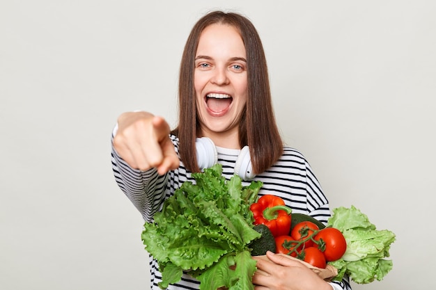 Photo excited brown haired young woman embraces bouquet of fresh vegetables wearing striped casual shirt pointing to camera asking you to keeps diet healthy eating isolated over gray background