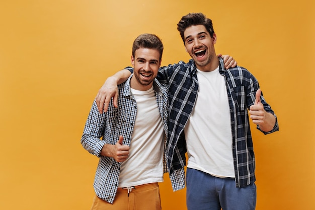 Excited brunet men in checkered shirts colorful pants and white tshirts smile show thumbs up and pose on orange background