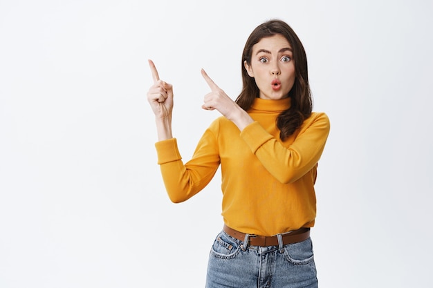 Excited brunette girl checking out promo offer, stare impressed at front, pointing at upper left corner banner, standing against white wall