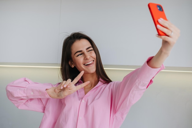 Excited brunette girl in pink shirt blinking making selfie at the kitchen blogger chatting