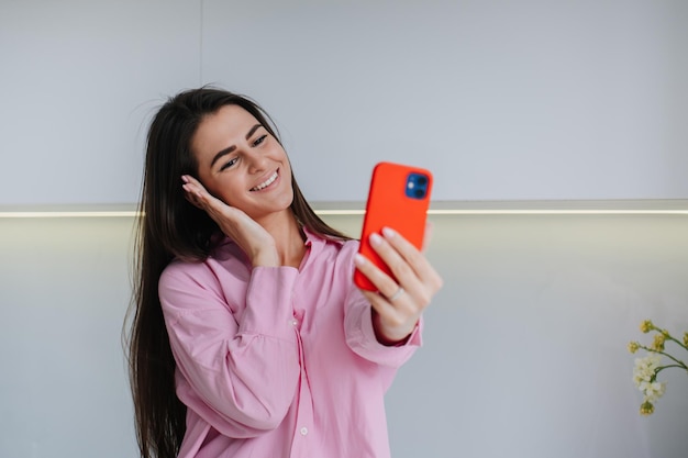 Photo excited brunette girl in pink shirt makes selfie by phone at kitchen blogger during travel tour