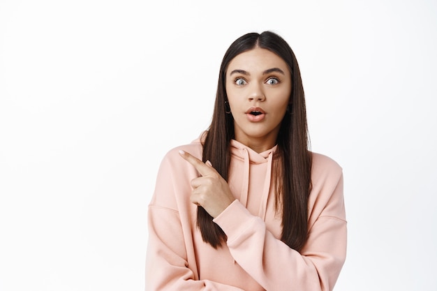 Excited brunette woman gasping and staring at front impressed, checking out awesome promo, pointing at upper left corner, showing banner logo on copy space