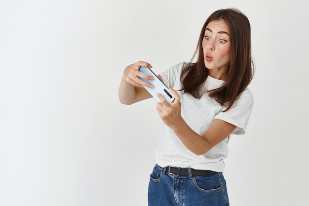 흥분한 브루네트 여성은 스마트폰으로 비디오 게임을 하는 동안 몸을 기울이고 즐겁게 보고 흰색 벽 위에 서 있습니다.