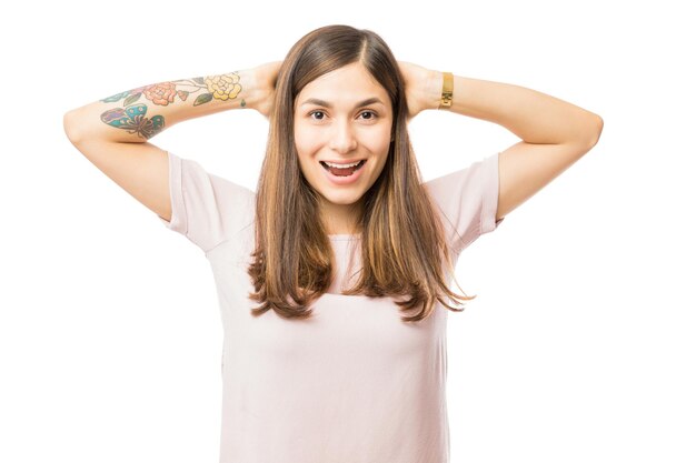 excited brunette woman with hands behind head standing isolated on white background