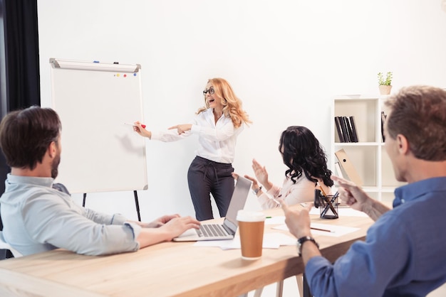 Excited businesswoman in eyeglasses pointing at blank whiteboard and looking at business partners