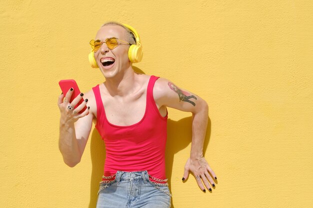 Взволнованный и беззаботный небинарный человек смеется и веселится, слушая музыку в наушниках и мобильном телефоне.