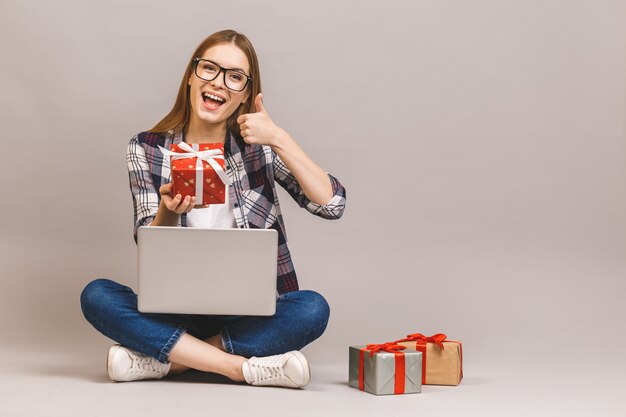 An excited casual girl holding laptop computer while sitting on a floor with stack of gift boxes. Thumbs up.