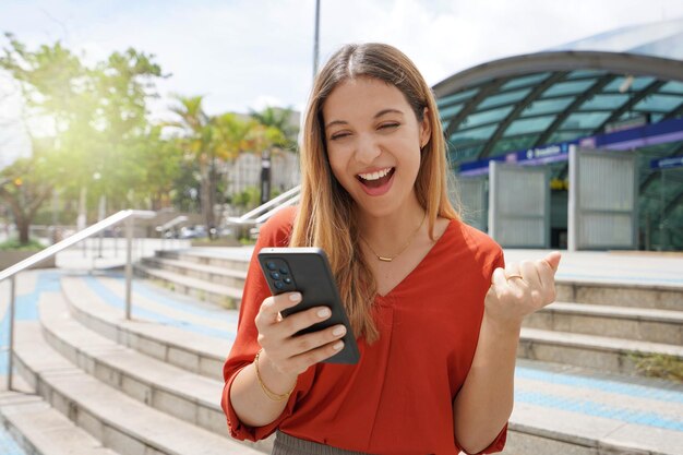 Взволнованная случайная женщина празднует хорошие новости на своем смартфоне в районе Бруклин в Сан-Паулу, Бразилия.