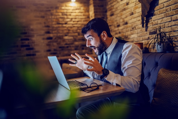 Excited caucasian bearded businessman in suit sitting in cafe and looking at laptop.