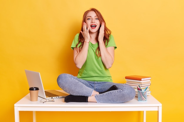 Excited Caucasian girl freelancer sitting on desk isolated on yellow wall. Achievement career. Education in university or college