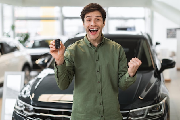 Foto uomo caucasico eccitato che tiene la chiave dall'auto che fa gesti di sì acquistando un'auto nuova di zecca presso la concessionaria