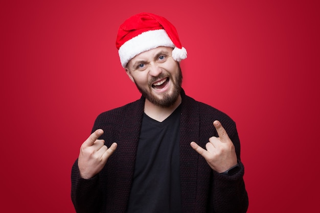 Excited caucasian man with santa hat gesturing rock and roll sign on a red studio wall