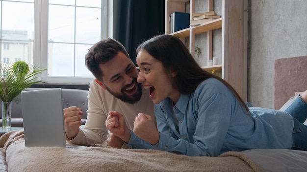 Excited caucasian married couple lying on bed at home win laptop achievement read amazing news