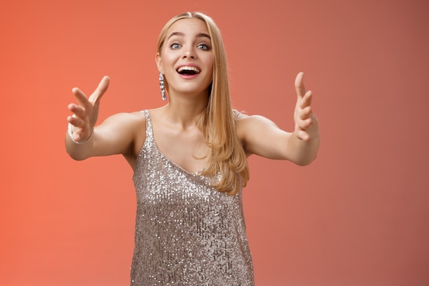 Photo excited charmign touched hearwarming young blond woman in silver party glamour dress stretch hands towards camera amused wanna hug cuddle hold cute puppy arms, standing red background.